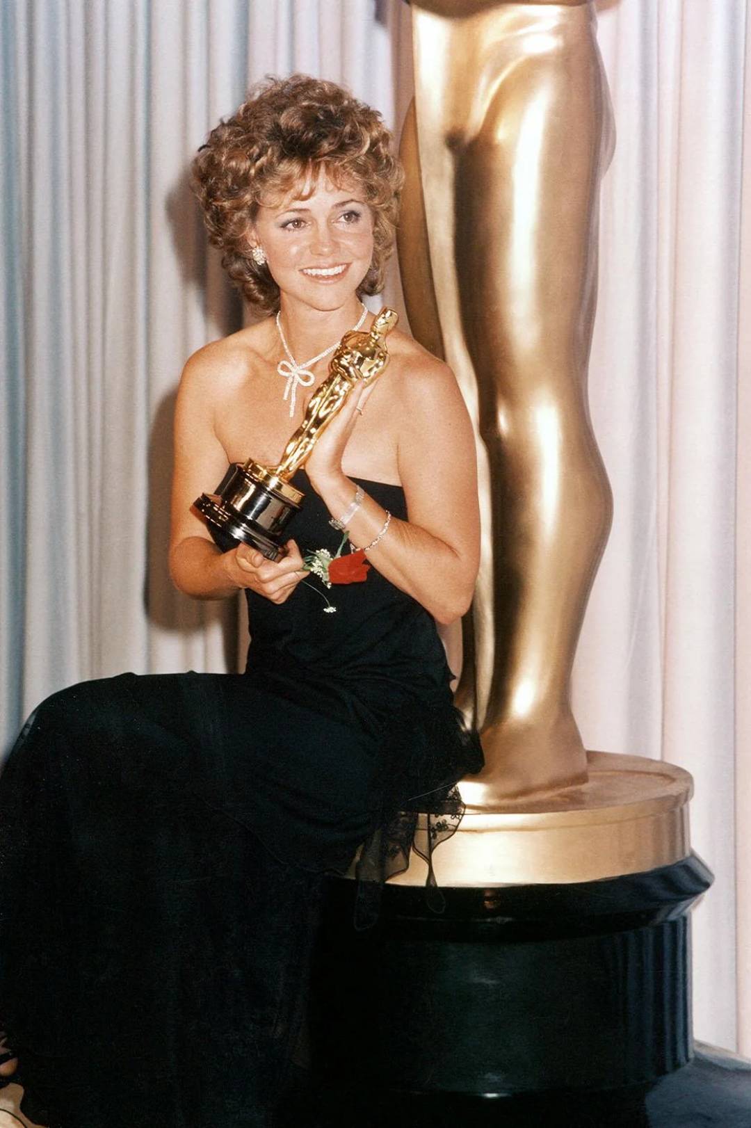 A woman in an elegant black dress sits smiling with an Oscar statuette in her hands, in front of a large golden Oscar statue and white curtains.