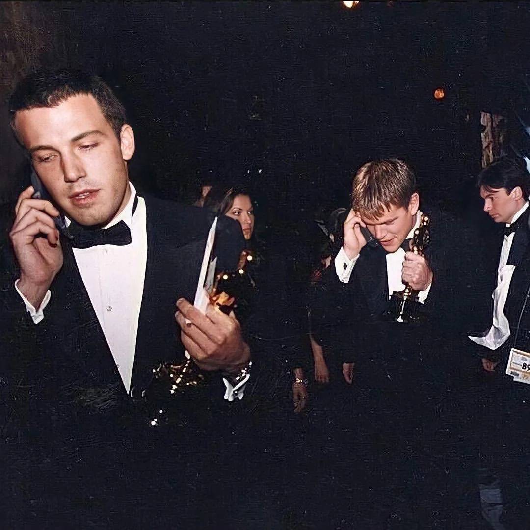 Two men in tuxedos hold Oscar statuettes and talk on cell phones at an event, with other attendees in the background. They appear to be backstage or in a hallway, celebrating their win.