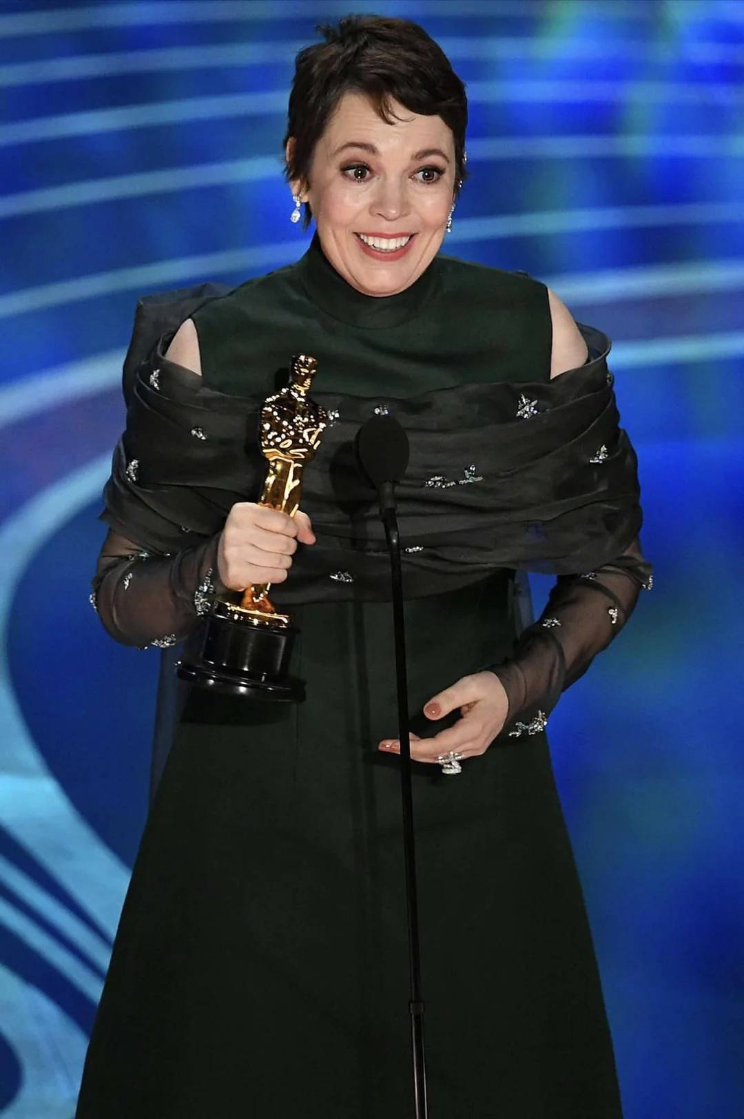 A woman in a dark green dress smiles while holding an Oscar trophy and standing at a microphone onstage, with a blue patterned background behind her.