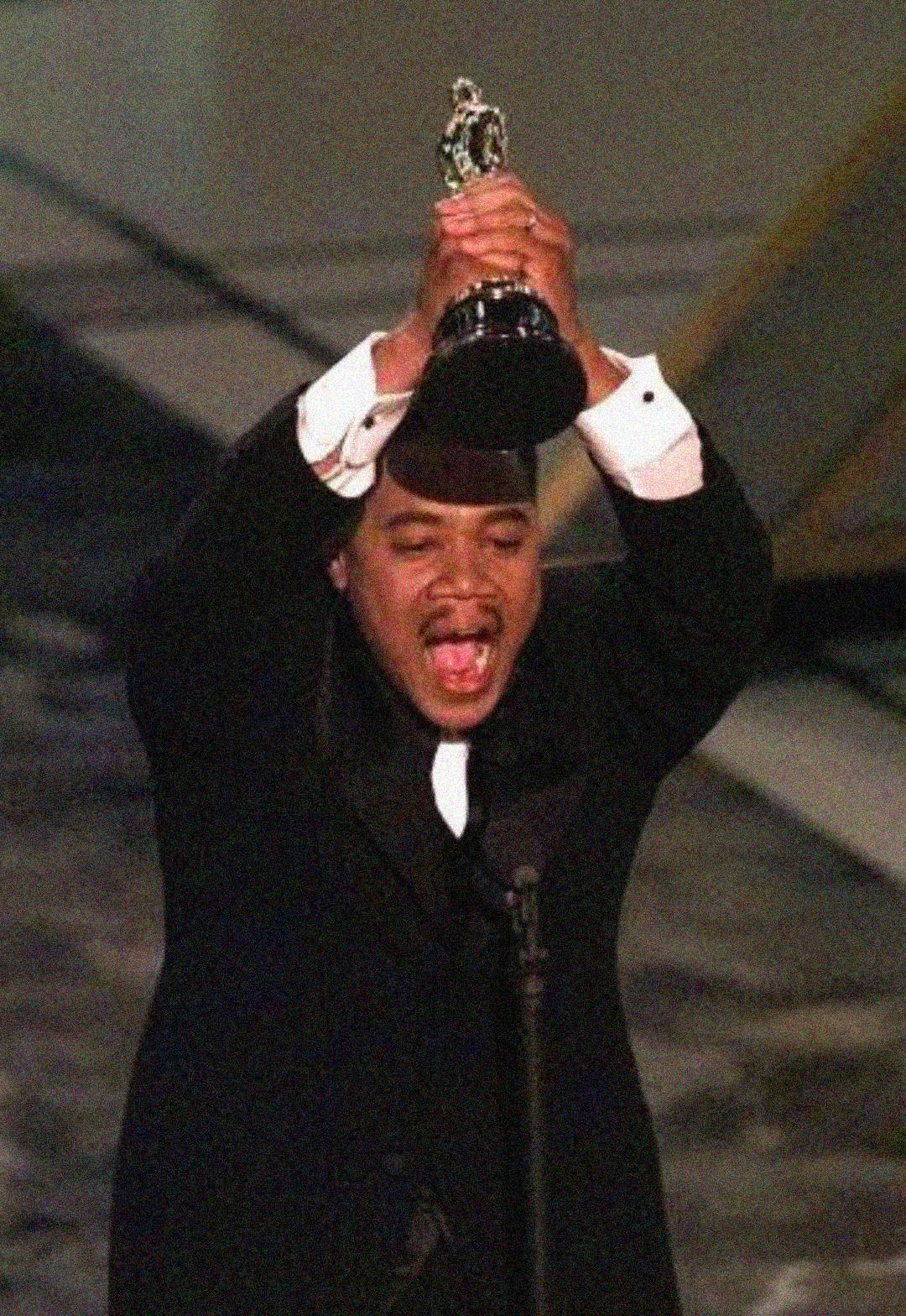 A man in a suit holds a trophy above his head and shouts joyfully on stage, appearing to celebrate winning an award.