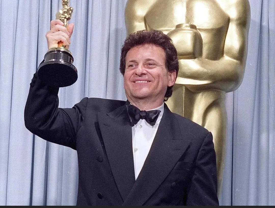 A man in a black tuxedo smiles and holds up an Oscar trophy. A large gold Oscar statue is visible in the background against blue curtains.