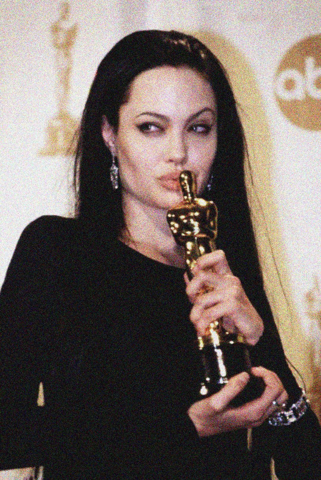 A woman with long dark hair in a black outfit holds and kisses an Oscar trophy while standing in front of a backdrop with gold Oscar statuettes and the ABC logo.
