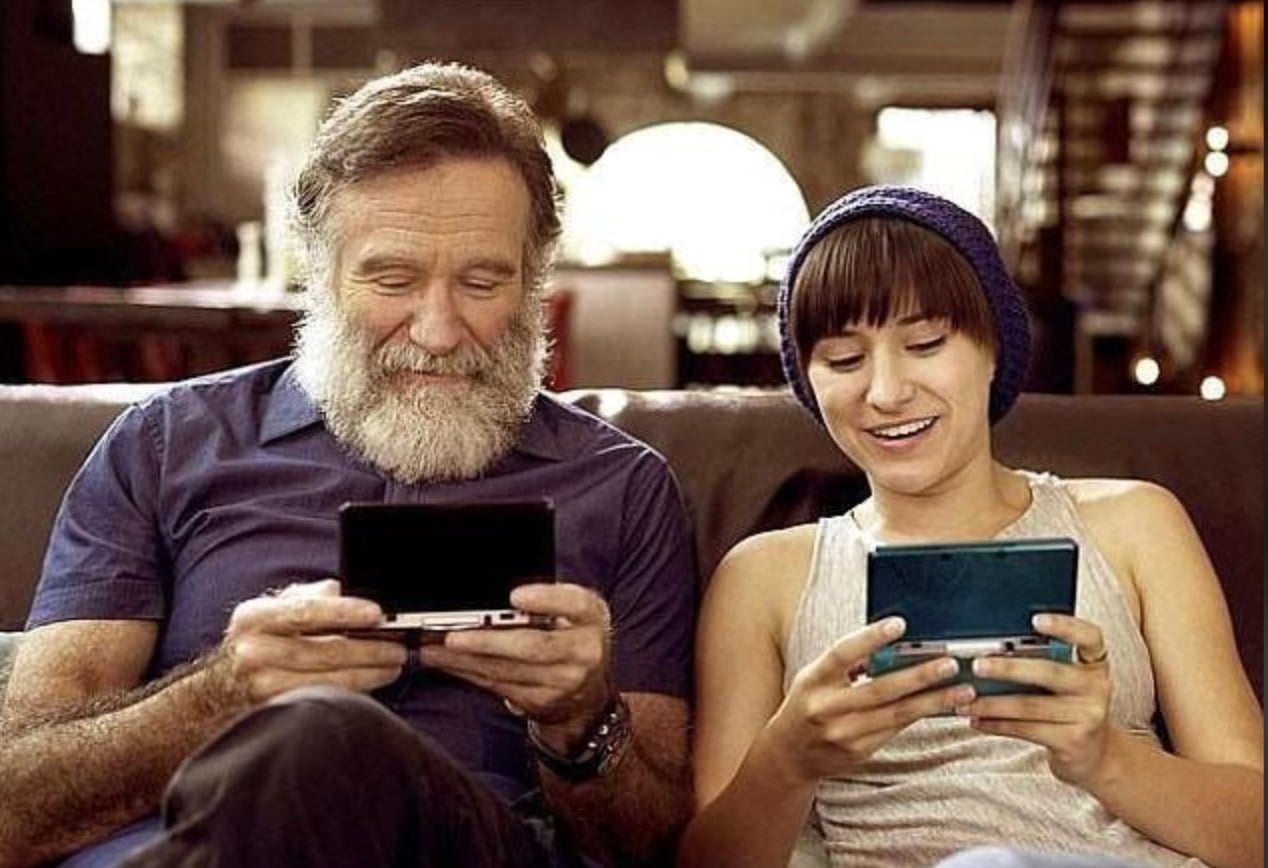 An older man with a beard and a younger person wearing a beanie sit on a couch, smiling and playing handheld gaming consoles together in a cozy, warmly lit room.