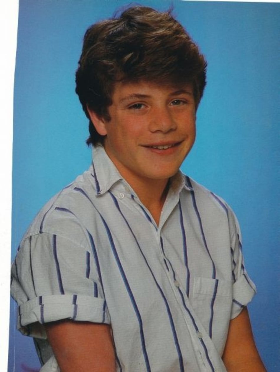 A smiling teenage boy with short brown hair wearing a white, short-sleeve, button-up shirt with blue stripes, sits in front of a blue background for a portrait-style photo.