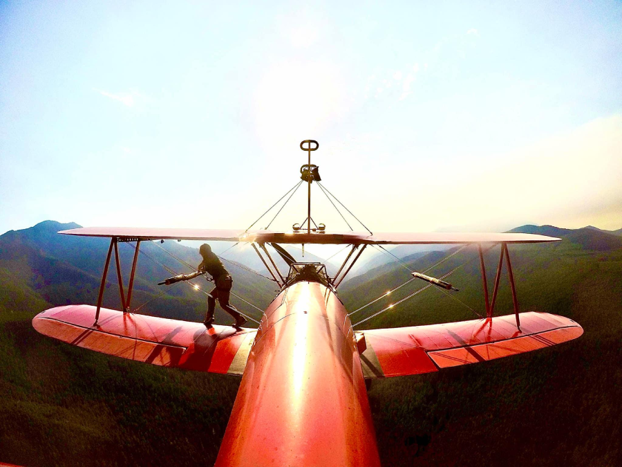 A person stands on the top wing of a red biplane flying over green hills and mountains, with the sun shining brightly in the background.