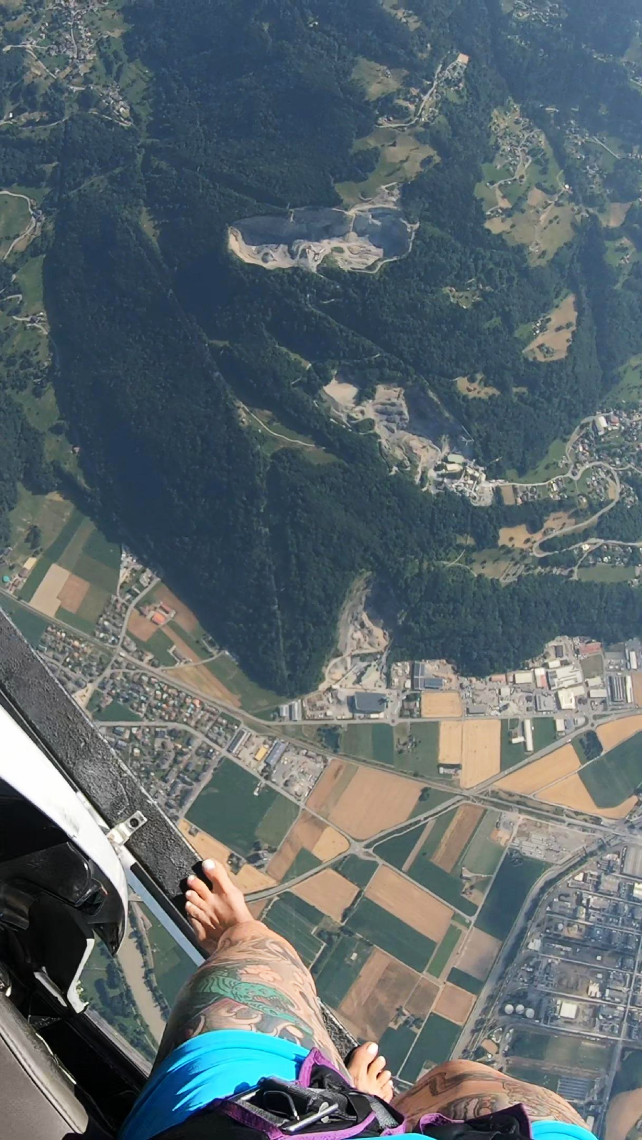 A person’s bare feet dangle out of an aircraft door high above a patchwork of fields, forests, and buildings, providing a dramatic aerial view of the landscape far below.
