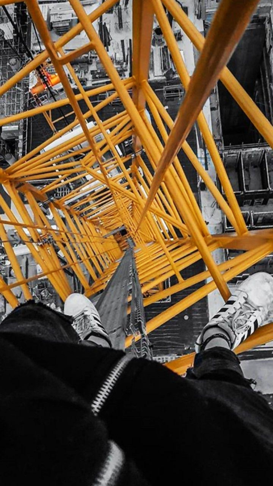 View looking down from high up on a yellow construction crane, with a person's feet visible at the edge. Buildings and construction materials are visible below. The image emphasizes height and perspective.