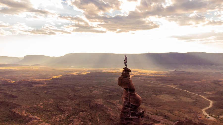 A person stands on top of a tall, narrow rock formation in a vast desert landscape at sunset, with light rays streaming through clouds and mountains in the background.