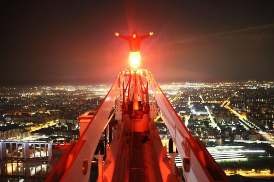A person stands with arms outstretched atop a tall crane at night, illuminated by red light, overlooking a brightly lit cityscape far below.
