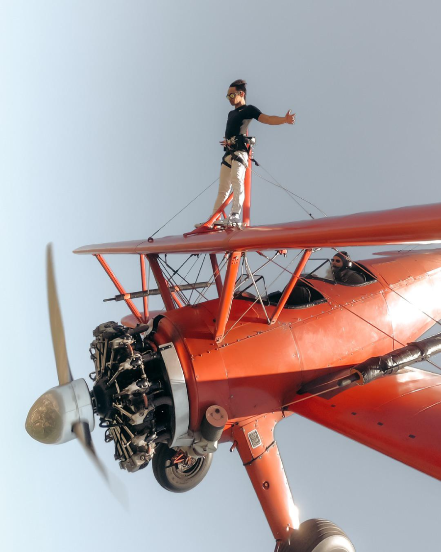A person stands on top of the wing of a red biplane in mid-flight, arms outstretched, wearing a harness and goggles, with the airplane's propeller spinning in the foreground against a clear sky.