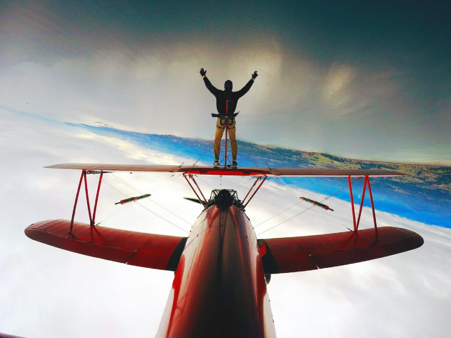 A person stands on top of a red biplane, arms raised, while the plane flies upside down over a landscape with clouds and water below.