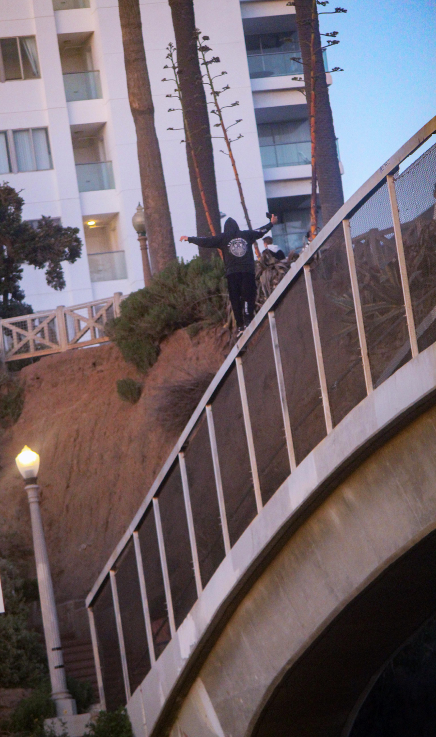 A person in a black hoodie walks on a narrow ledge above a curved bridge railing, arms outstretched for balance, with tall buildings, trees, and a streetlamp in the background.