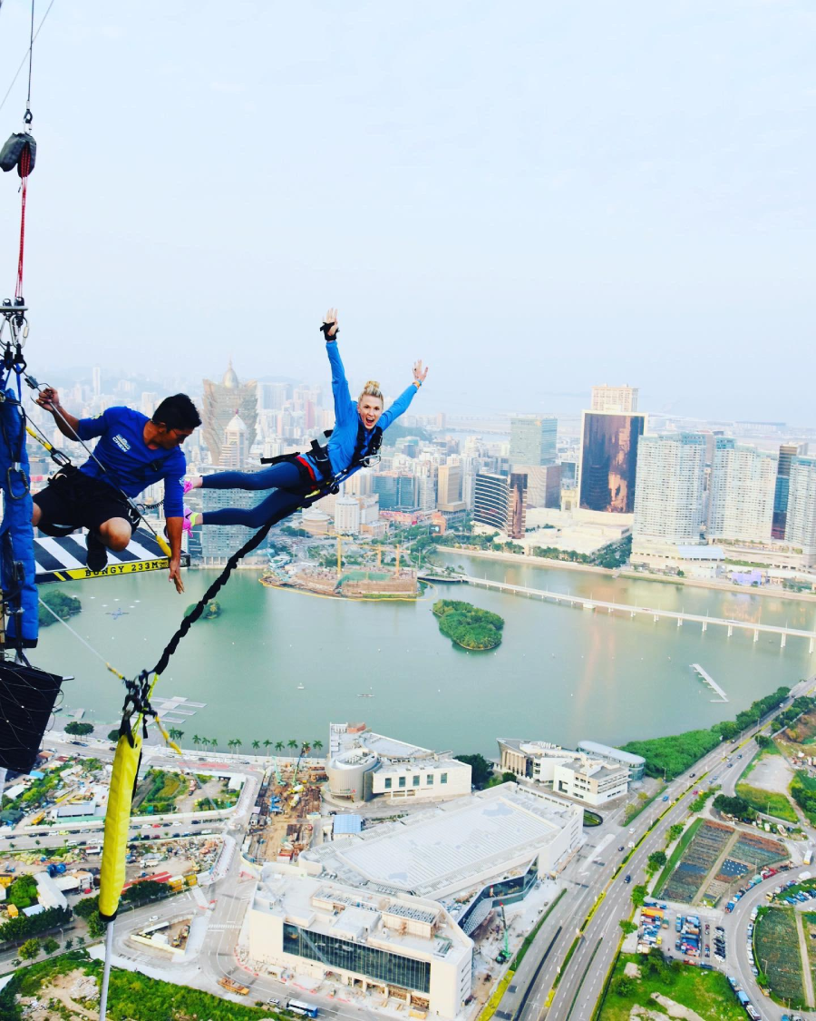 A person is bungee jumping off a tall platform with a guide assisting, arms raised in excitement, above a city skyline with tall buildings, a river, and roads visible below.