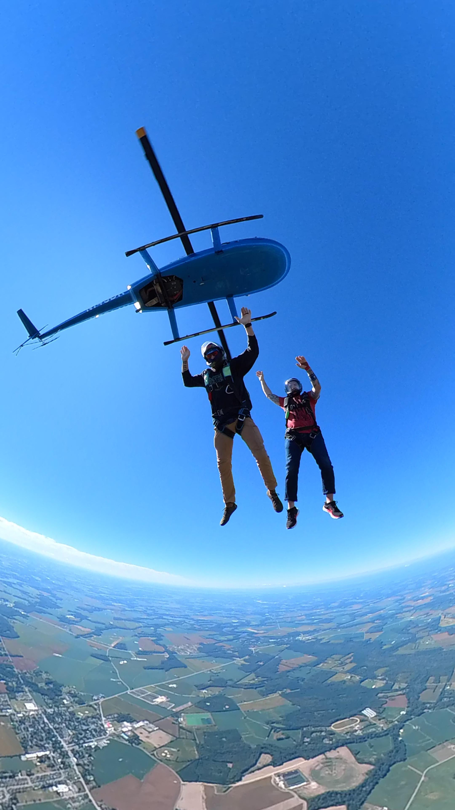 Two skydivers in midair after jumping from a blue helicopter, with clear blue sky above and farmland visible far below. Both are wearing helmets and harnesses, free-falling with arms raised.