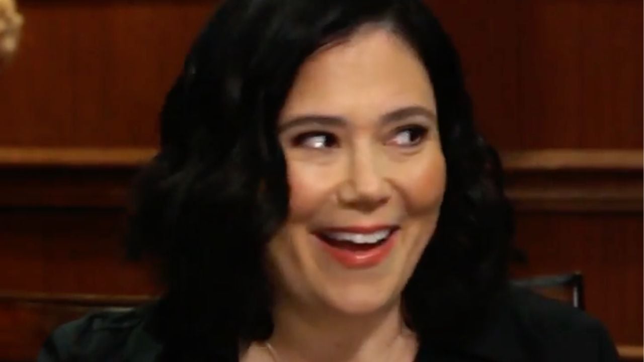 A woman with wavy dark hair smiles and looks to her left. She is indoors with a wooden background.