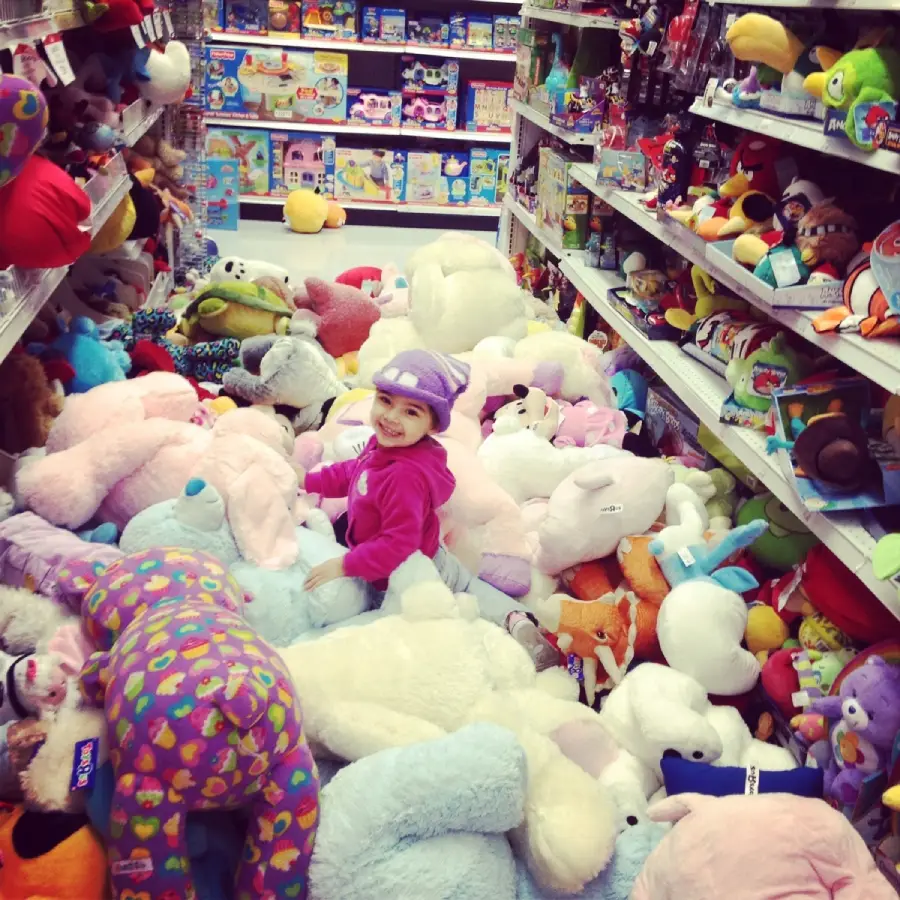 A young child in a purple outfit and hat smiles while sitting among a large pile of colorful stuffed animals in a toy store aisle, surrounded by shelves filled with toys and plushies.
