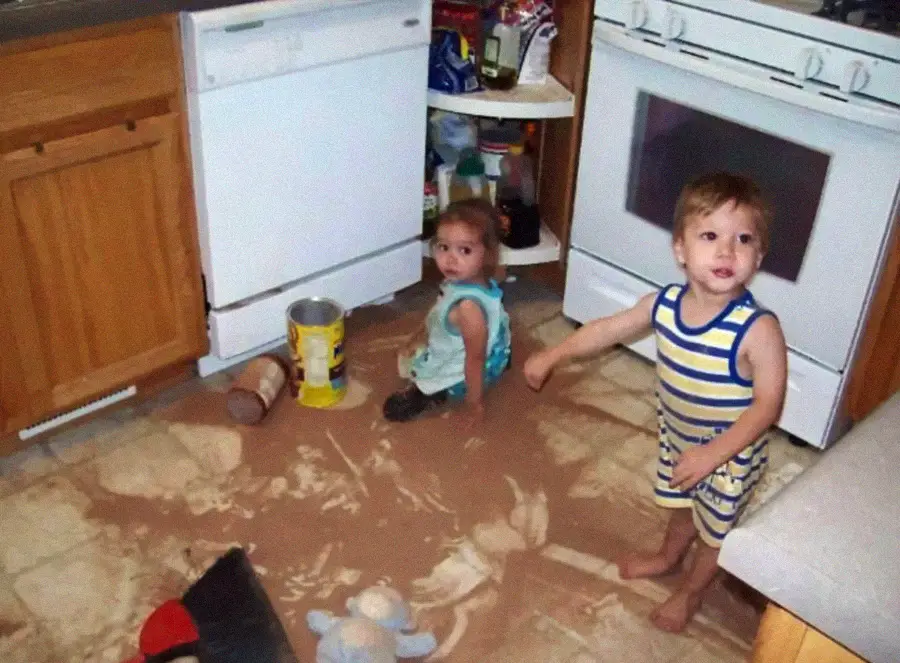 Two young children in a kitchen are surrounded by spilled powder on the floor, with open cabinets and a tipped-over canister nearby. Both kids look up, one sitting and one standing, appearing surprised or curious.