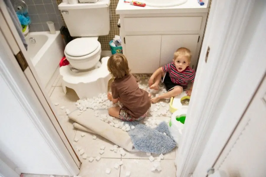 Two young children sit on a bathroom floor covered in pieces of white packing peanuts, with a rug pushed aside and a surprised expression on one child's face. The bathroom looks messy and playful.
