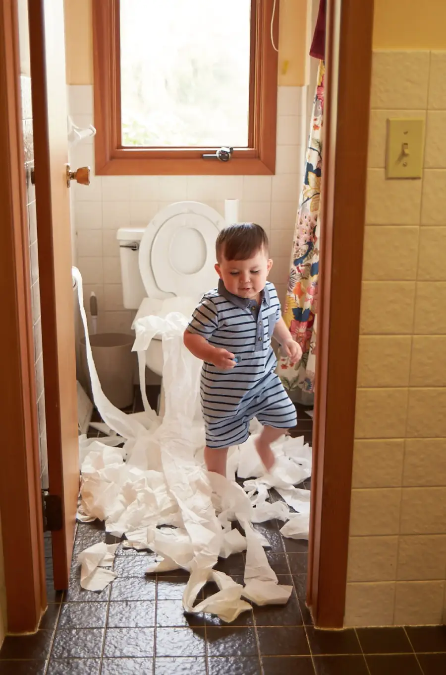 A toddler in striped pajamas stands in a bathroom with toilet paper strewn all over the floor and hanging from the toilet. The room is lit by natural light coming through a window behind the toilet.
