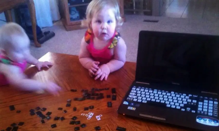 Two young children in pink outfits sit at a wooden table, surrounded by loose keyboard keys from a laptop. The laptop is open to the right, while one child looks up and the other reaches toward the scattered keys.
