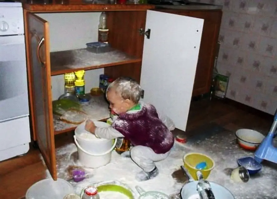 A young child sits on the kitchen floor, covered in flour, surrounded by scattered bowls, dishes, and a bucket. An open cabinet reveals more items, and flour is spilled everywhere. The child appears to be playing with the mess.