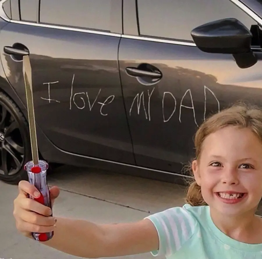 A smiling young girl holds a screwdriver in front of a black car, which has "I love my dad" scratched into the door.