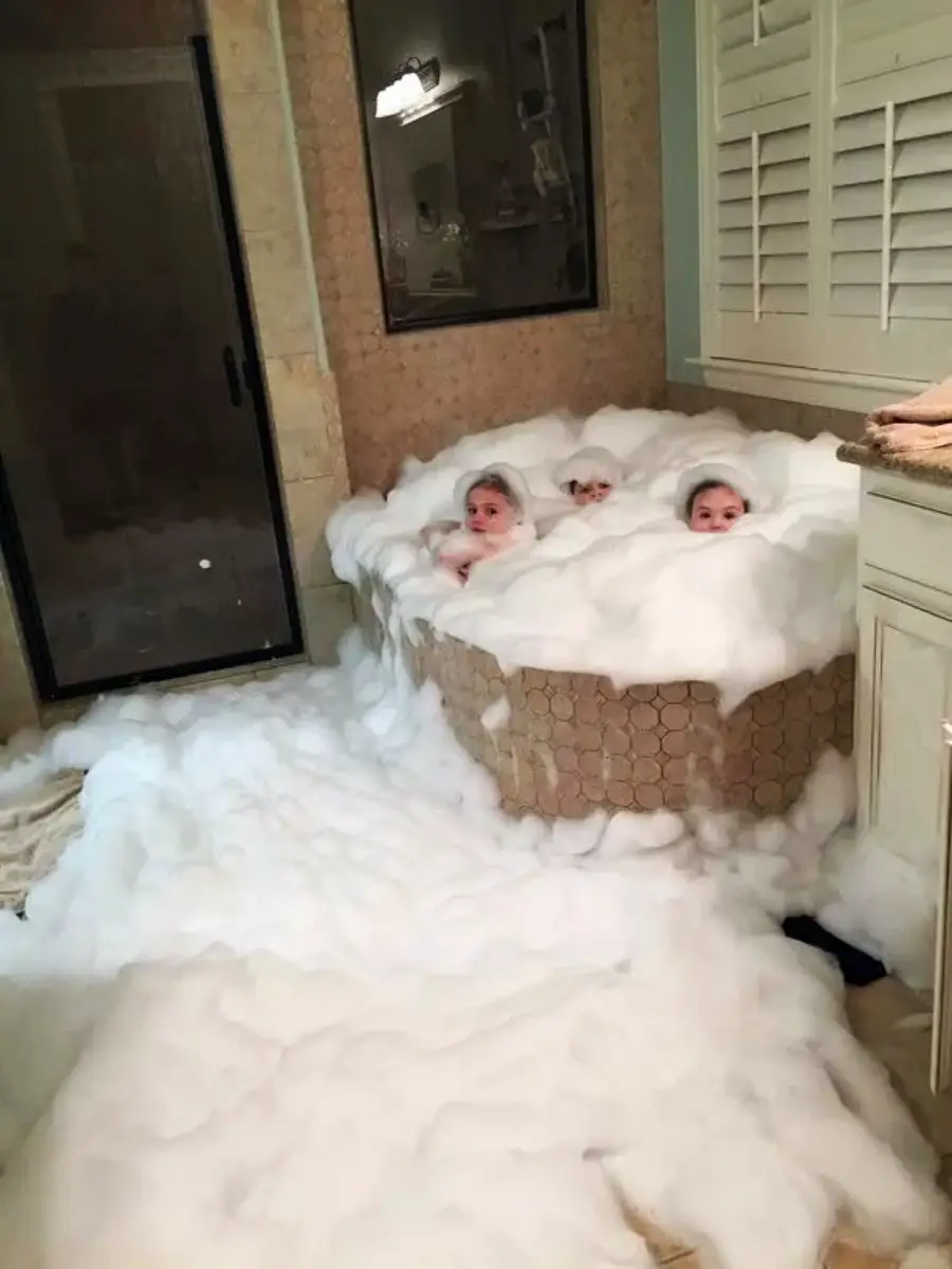 Three children sit in a bathtub overflowing with white bubbles, which spill onto the bathroom floor, nearly covering everything. Only their faces are visible above the foam.