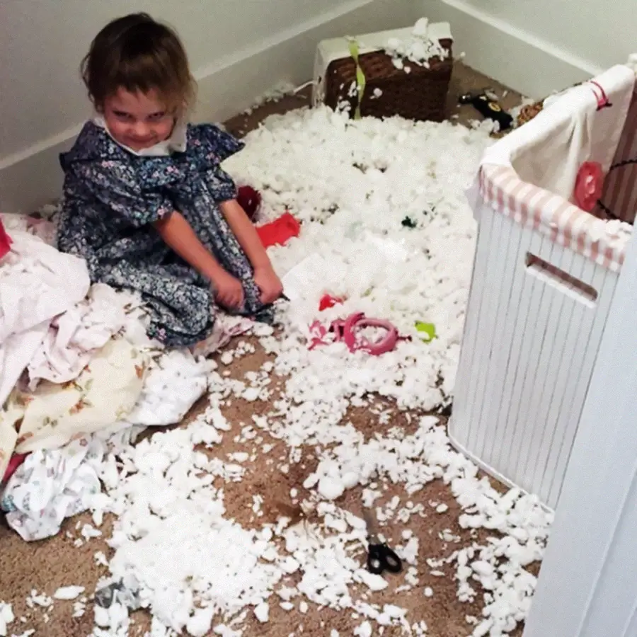 A young child in a dress sits on a bedroom floor covered with scattered white stuffing, likely from a torn-up pillow. The child smiles mischievously amid the mess, with laundry baskets and blankets nearby.