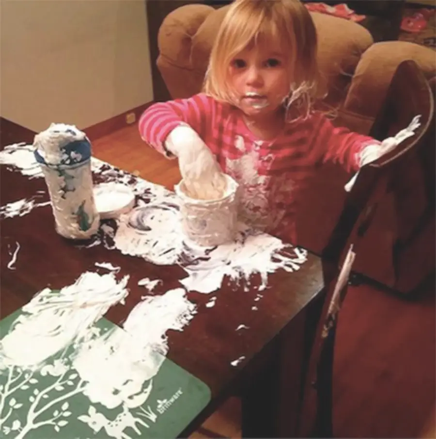 A young child with blonde hair and a pink striped shirt is covered in white cream, sitting at a table with messy cream smeared on her hands, face, and all over the table and chair.
