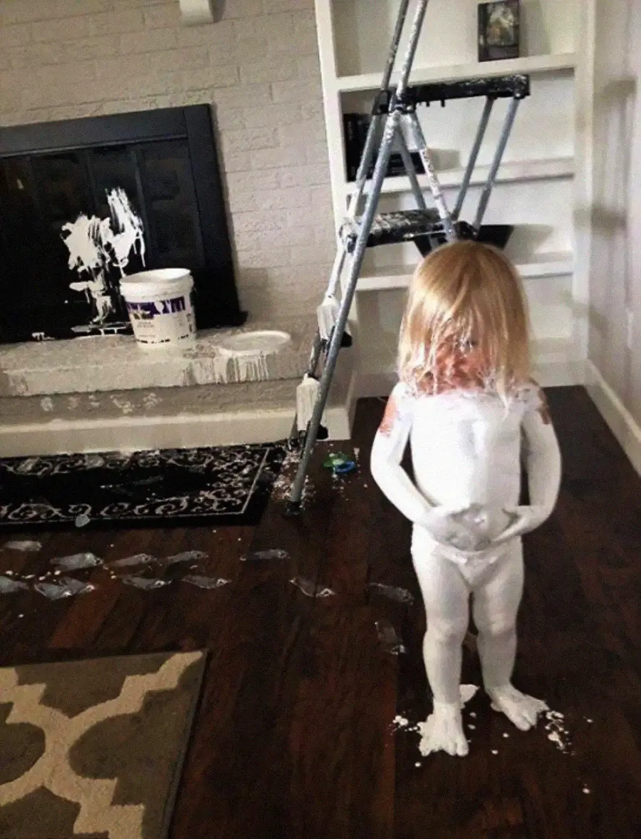 A young child stands covered in white paint, with paint splattered on the floor, ladder, fireplace, and nearby surfaces, suggesting a playful or messy accident during painting.