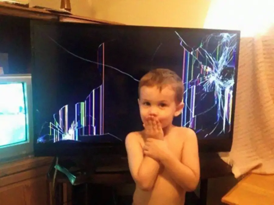 A young child stands shirtless in front of a large TV with a severely cracked and colorful broken screen, covering their mouth with both hands and looking at the camera.