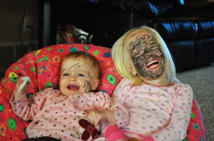 Two young children in pink pajamas sit on a colorful beanbag, smiling. Both have scribbles of black marker all over their faces, suggesting playful mischief.