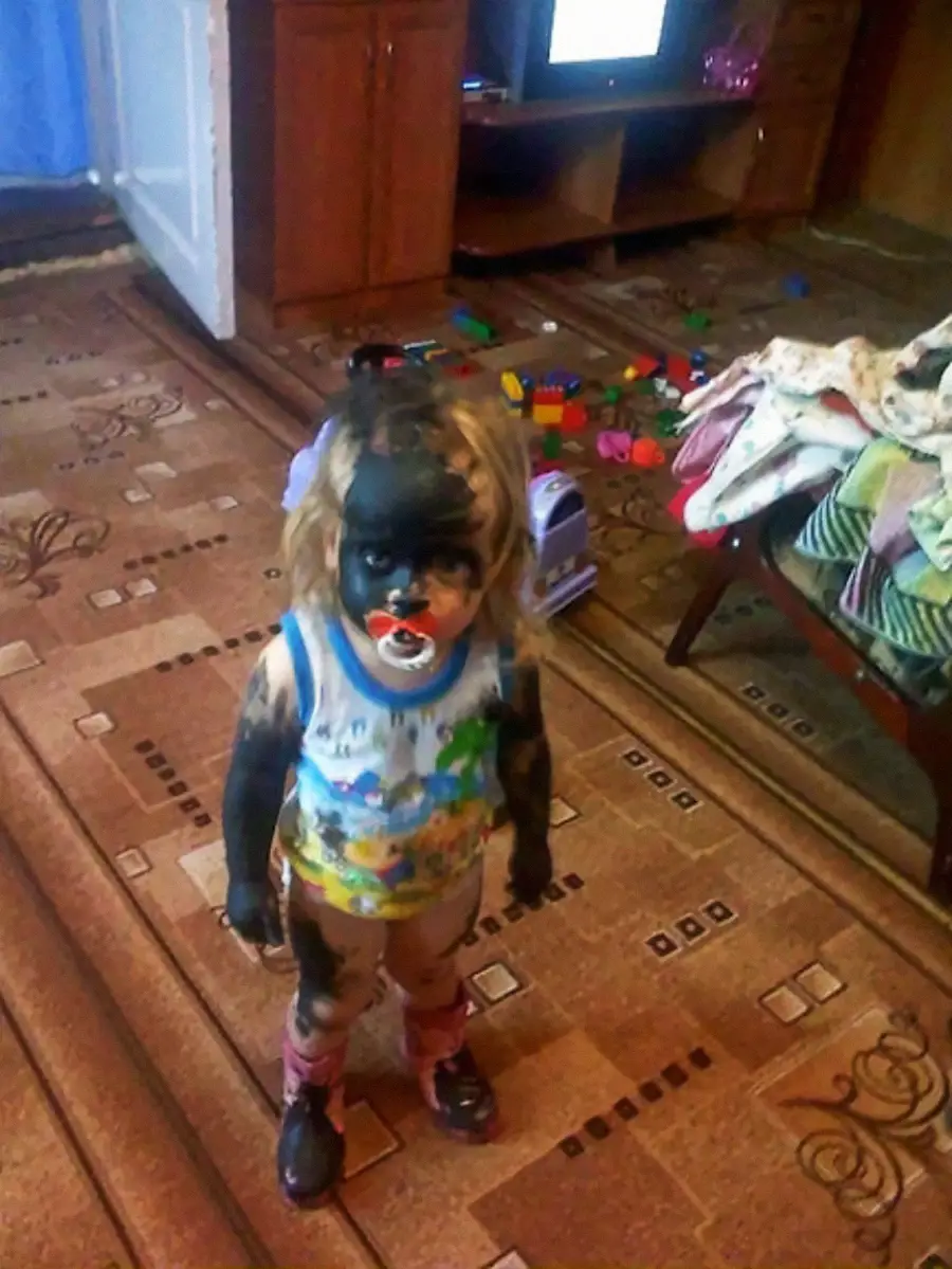 A young child stands indoors with their face, arms, and legs covered in black marker or paint, wearing a colorful shirt, pink socks, and a pacifier, surrounded by toys and household furniture.