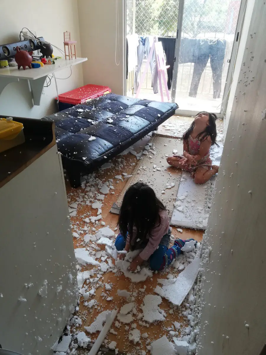Two young children sit on the floor of a messy room, surrounded by torn pieces of white foam scattered everywhere. The black futon and the rest of the room are covered in foam bits. A balcony with laundry is visible in the background.