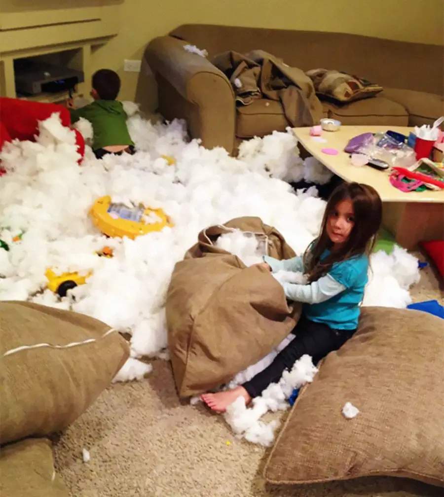 Two children sit among a huge mess of white stuffing pulled from cushions, which covers the floor and furniture in a living room. Toys and household items are scattered around the chaotic scene.