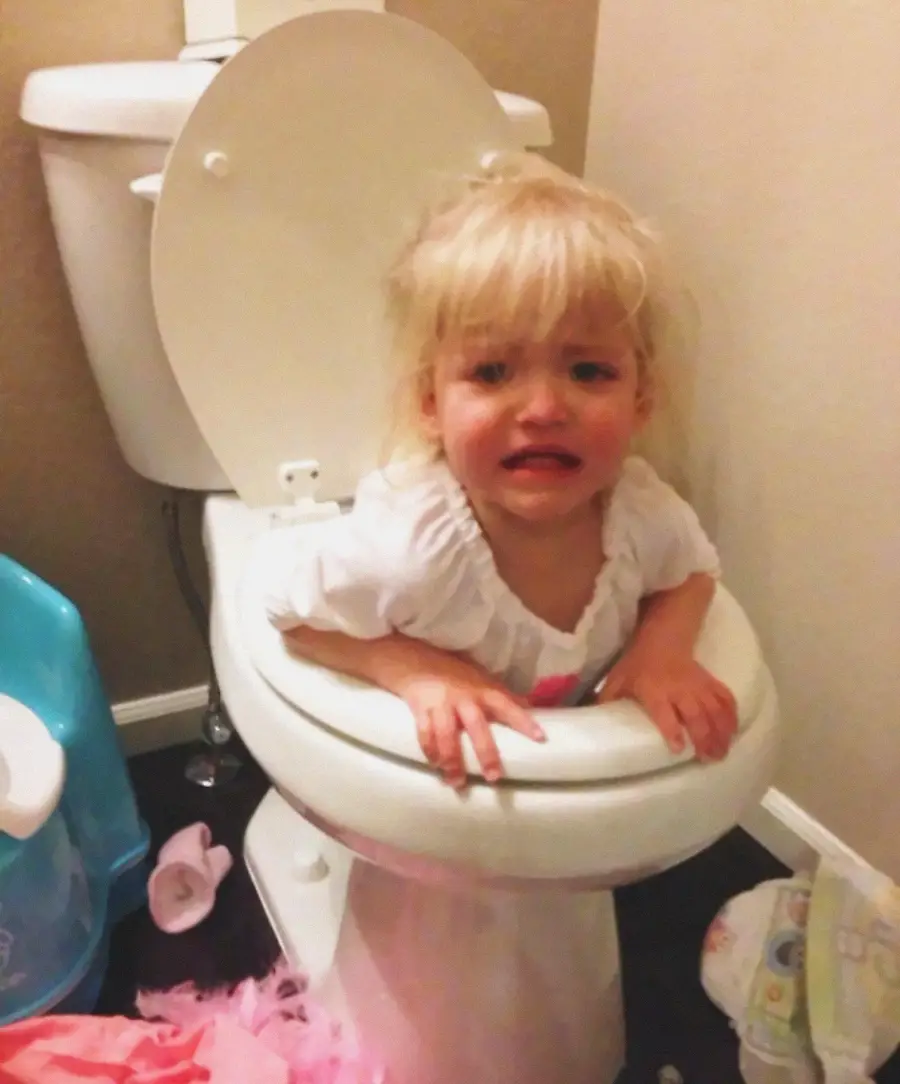 A young child with blonde hair and a white shirt is stuck sitting inside a toilet bowl, looking upset and crying in a bathroom. Nearby, there are scattered clothes and a potty seat on the floor.