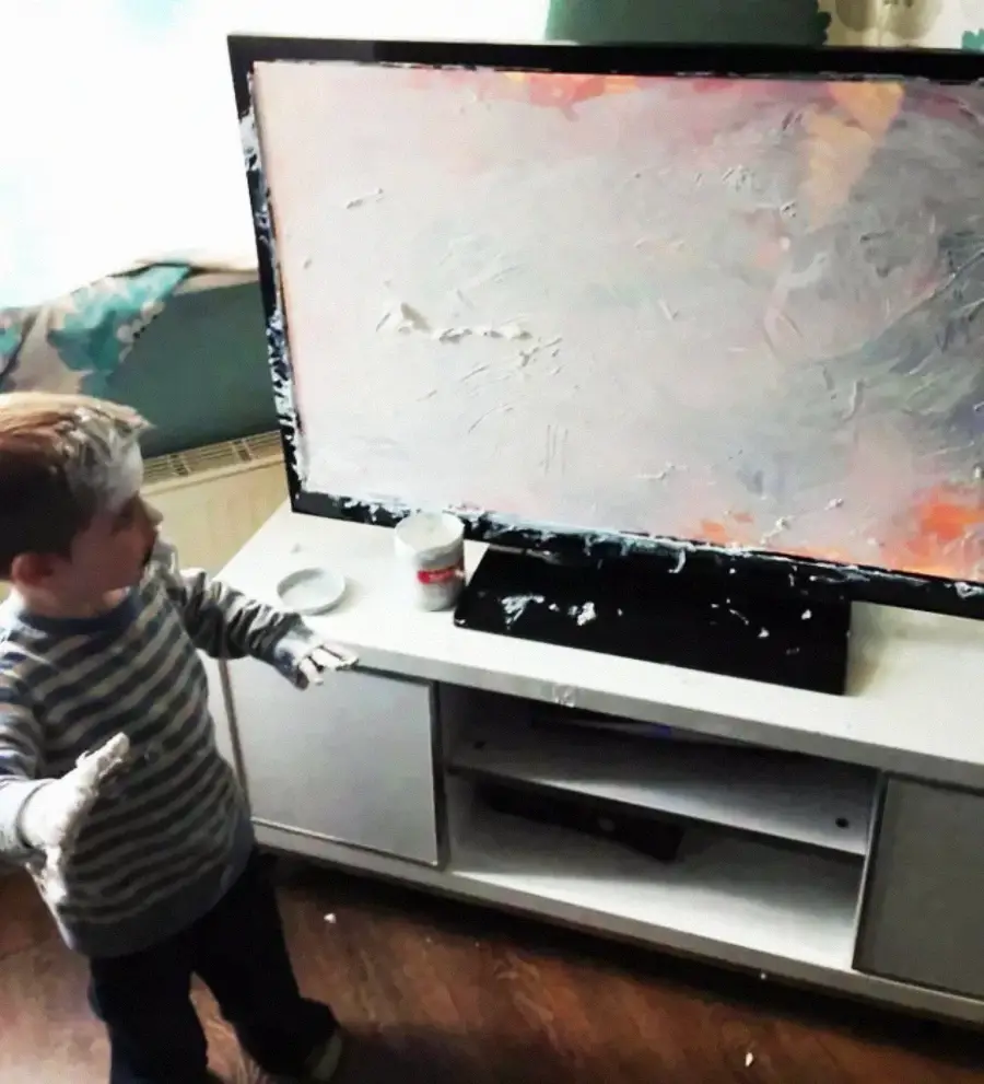 A young child with white cream or paint on their hands stands beside a TV that has been smeared with the same substance, with a jar and the TV remote on the cabinet nearby.