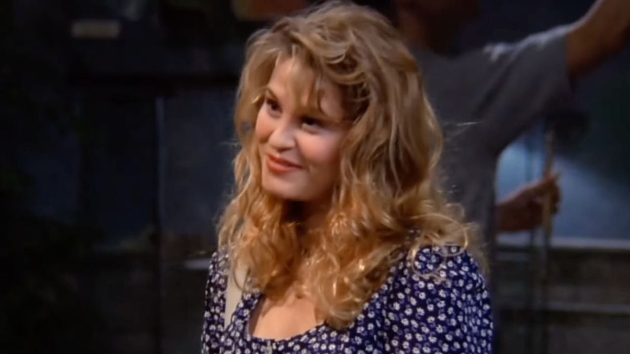 A woman with wavy blonde hair wearing a blue patterned dress smiles softly, looking slightly to the side. The background is dimly lit, with a blurred figure standing behind her.