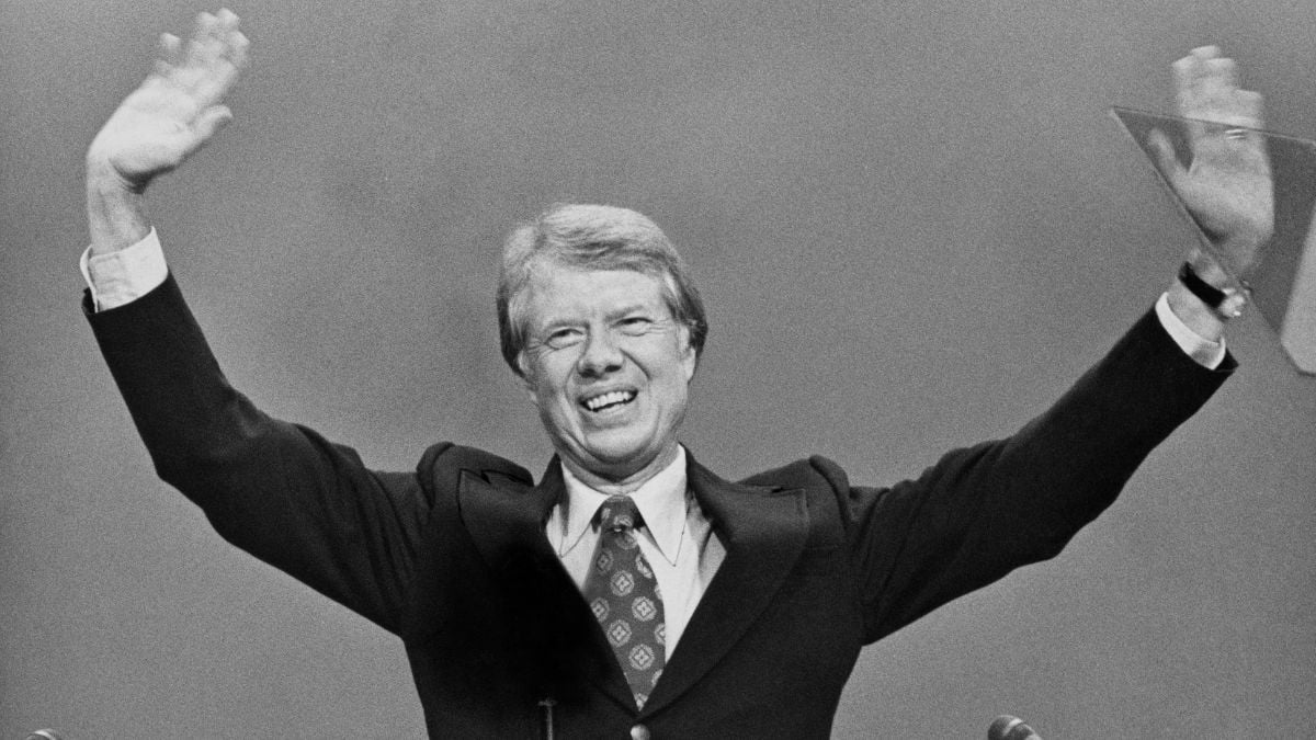 A man in a suit and tie smiles and raises both arms high, waving to an audience, standing in front of a plain background. Black and white photo.