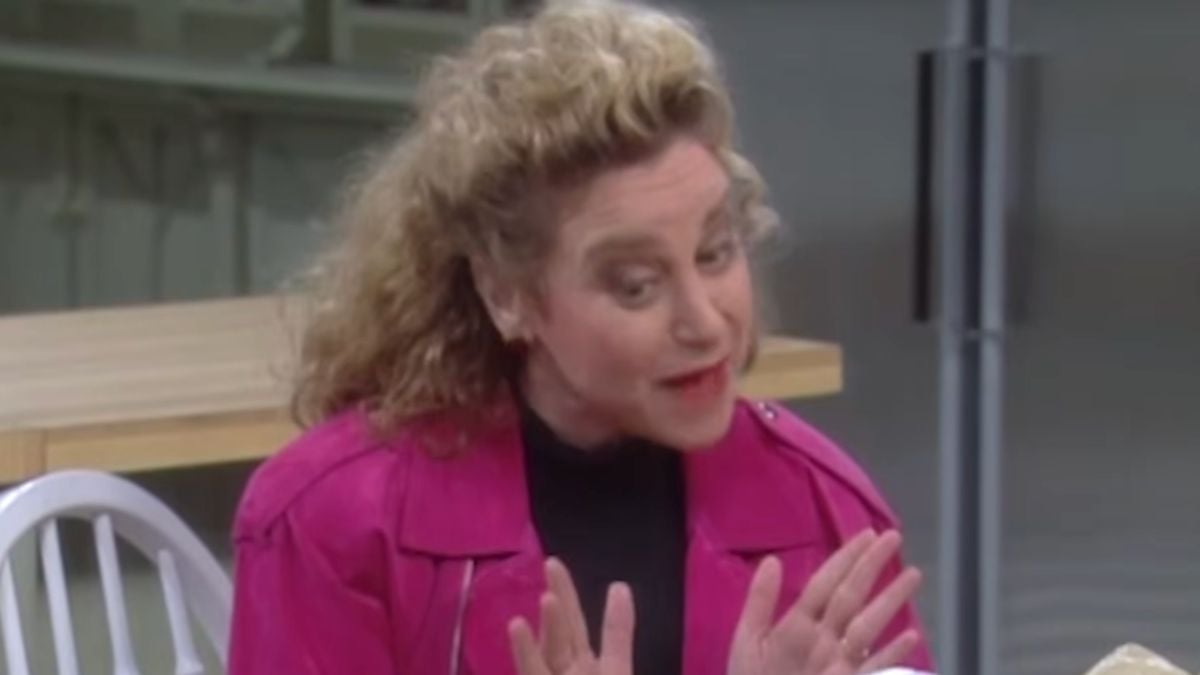 A woman with curly blonde hair, wearing a bright pink jacket and a black top, gestures with her hands while speaking, sitting at a table with a white chair behind her.