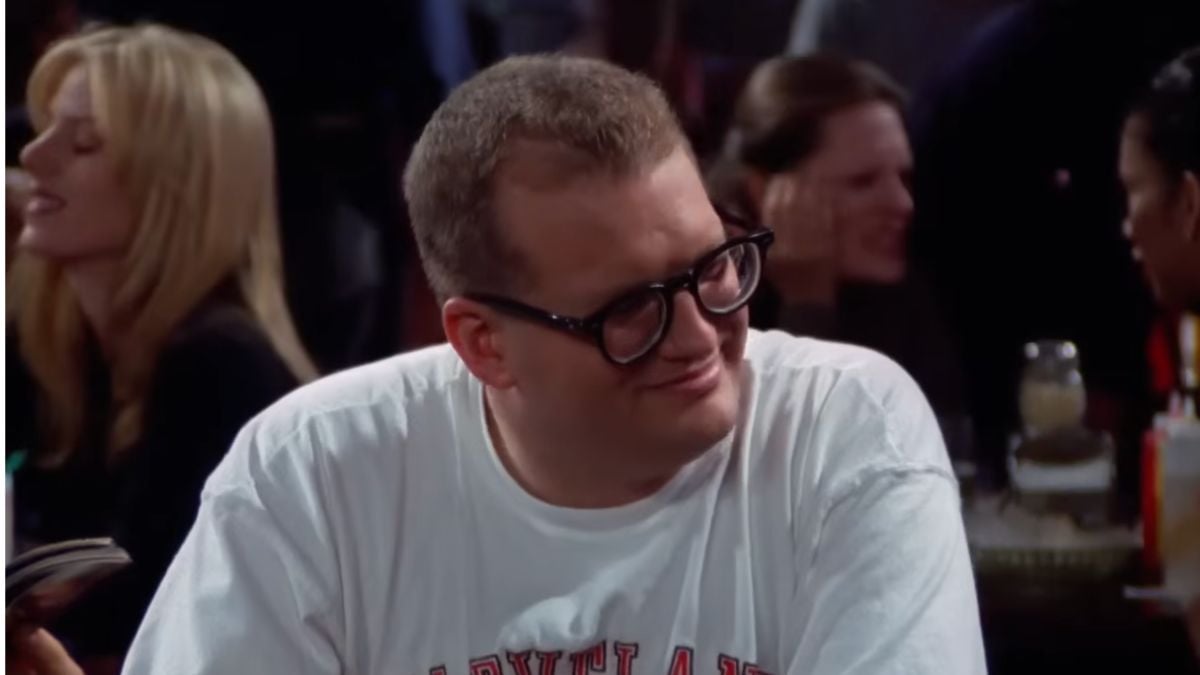 A man with glasses and a white shirt sits at a table in a crowded indoor setting, smiling slightly. People are talking in the blurred background.
