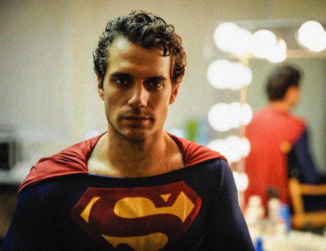 A man with curly hair wearing a Superman costume stands in a dressing room, facing the camera. Bright lights and a mirror with his reflection are visible in the background.