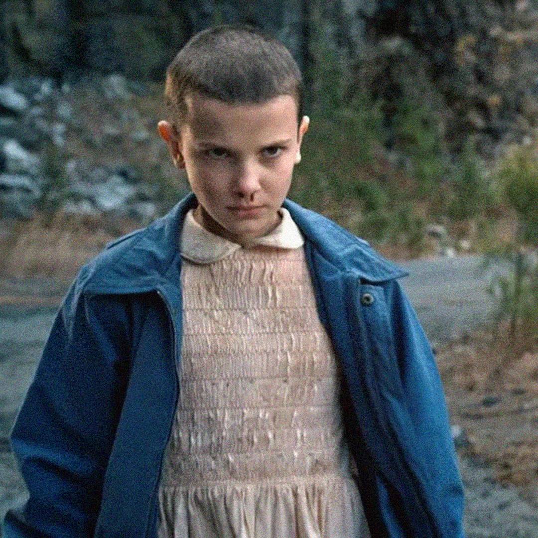 A young person with short hair wears a pale dress and a blue jacket, standing outdoors on a dirt path. They have a serious, intense facial expression and are looking forward. Trees and rocks are visible in the background.