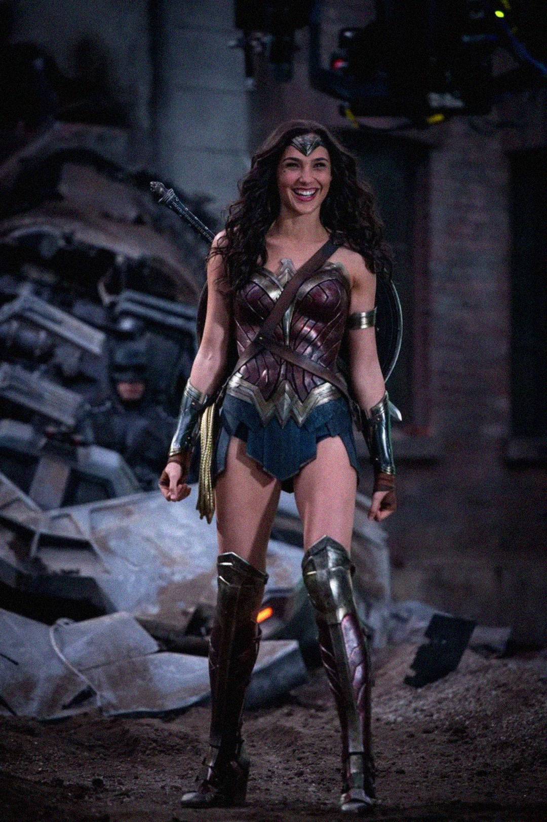 A woman dressed as Wonder Woman stands smiling in a battle-damaged urban setting, wearing armor, a tiara, and boots, with a sword and shield on her back.