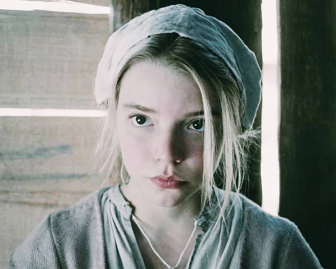 A young woman with pale skin and light hair, wearing a white headscarf and simple, old-fashioned clothing, looks seriously at the camera inside a wooden structure with sunlight filtering through the gaps.