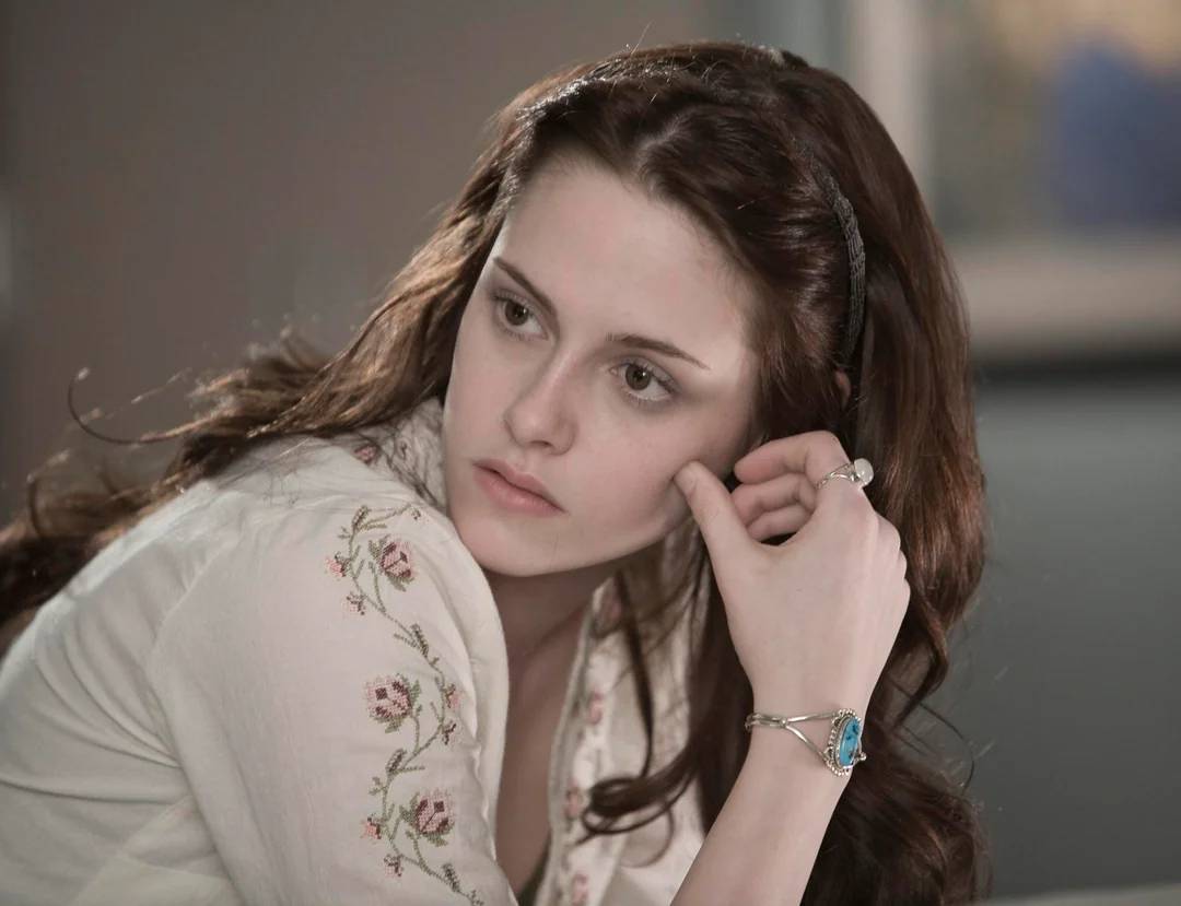 A young woman with long brown hair, wearing a white embroidered shirt and a silver bracelet with a blue stone, rests her chin on her hand and looks to the side with a thoughtful expression.