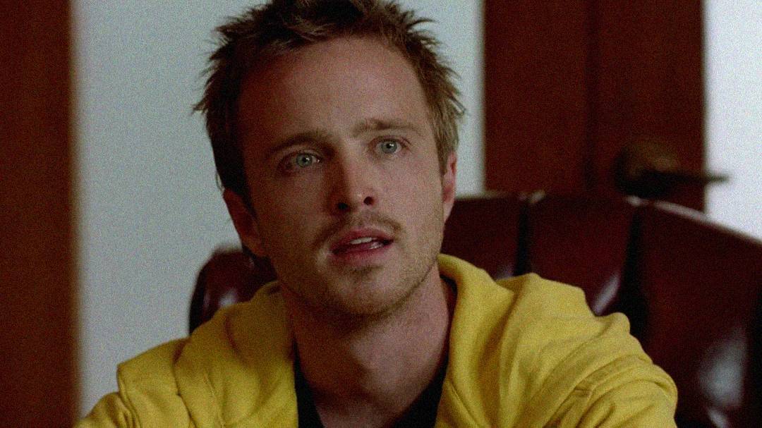 A young man with short messy hair and light facial hair, wearing a yellow hoodie, sits indoors and looks slightly upward with a concerned or thoughtful expression.