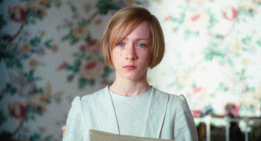 A young girl with short blonde hair and blue eyes stands indoors, holding a piece of paper. She wears a light-colored dress, and behind her is floral-patterned wallpaper.