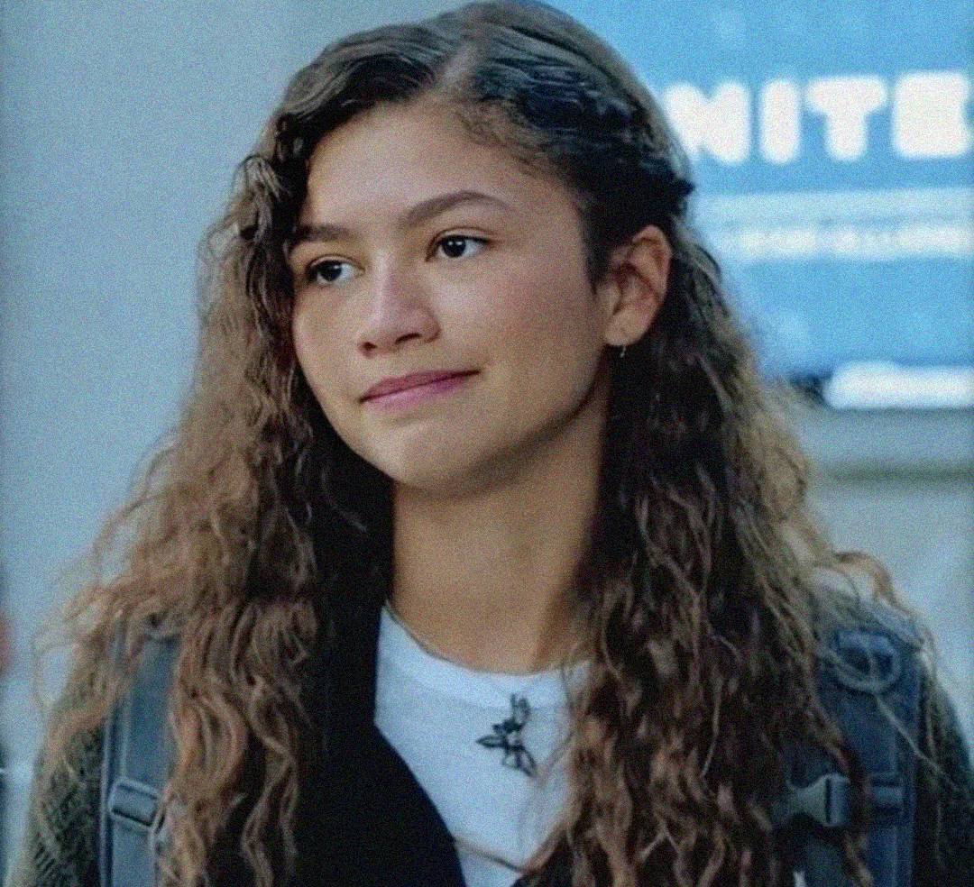 A young woman with long, curly brown hair stands outside, wearing a backpack and a white t-shirt under a dark jacket. She has a slight smile and looks ahead. The background is blurred with a blue and white sign partially visible.