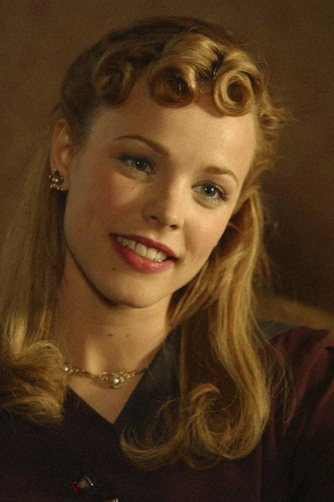 A young woman with vintage-styled blonde hair, pearl earrings, and a necklace smiles warmly. She is wearing dark clothing and is set against a soft brown background.